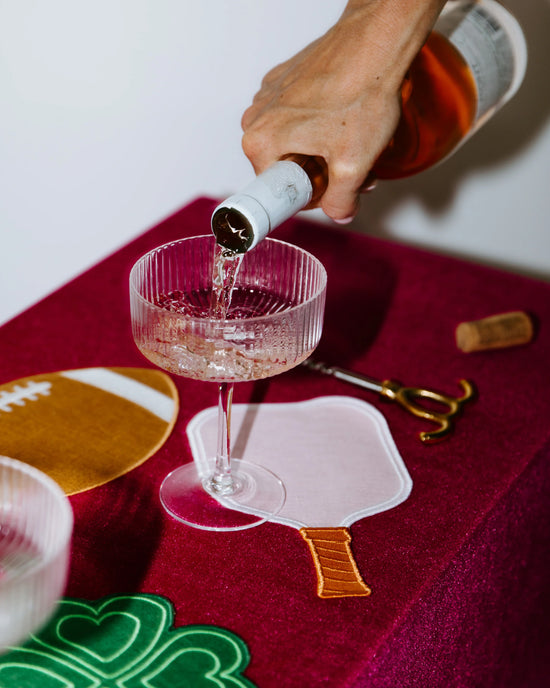 Person pouring champagne into a glass, set on a red table with a football coaster and decorative items.