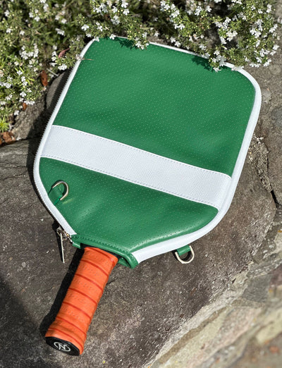 Green and white pickleball paddle with an orange grip, resting on a stone surface near greenery.