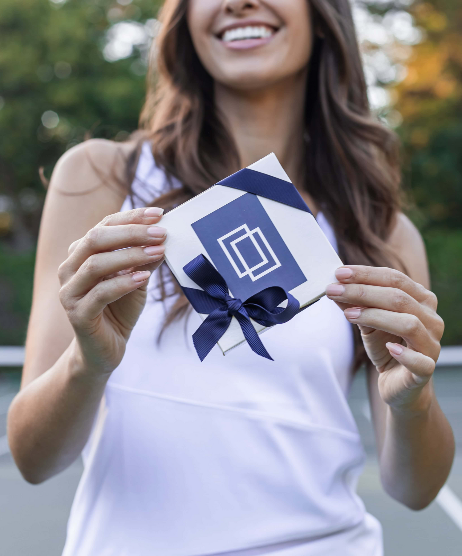 Smiling woman in a white tank top holding a gift box with a blue ribbon outdoors.