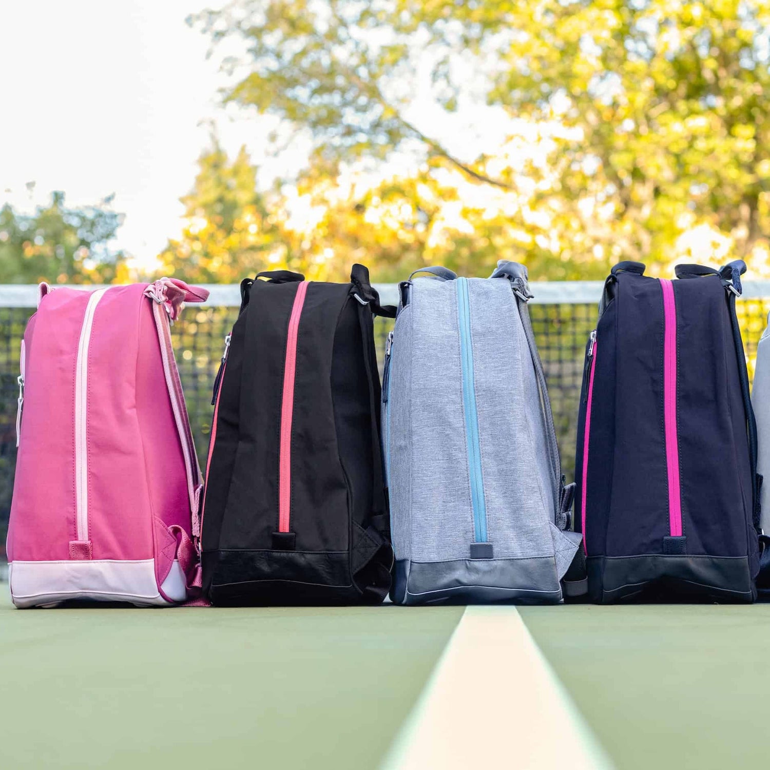 Four colorful sports bags on a tennis court with a net and trees in the background.