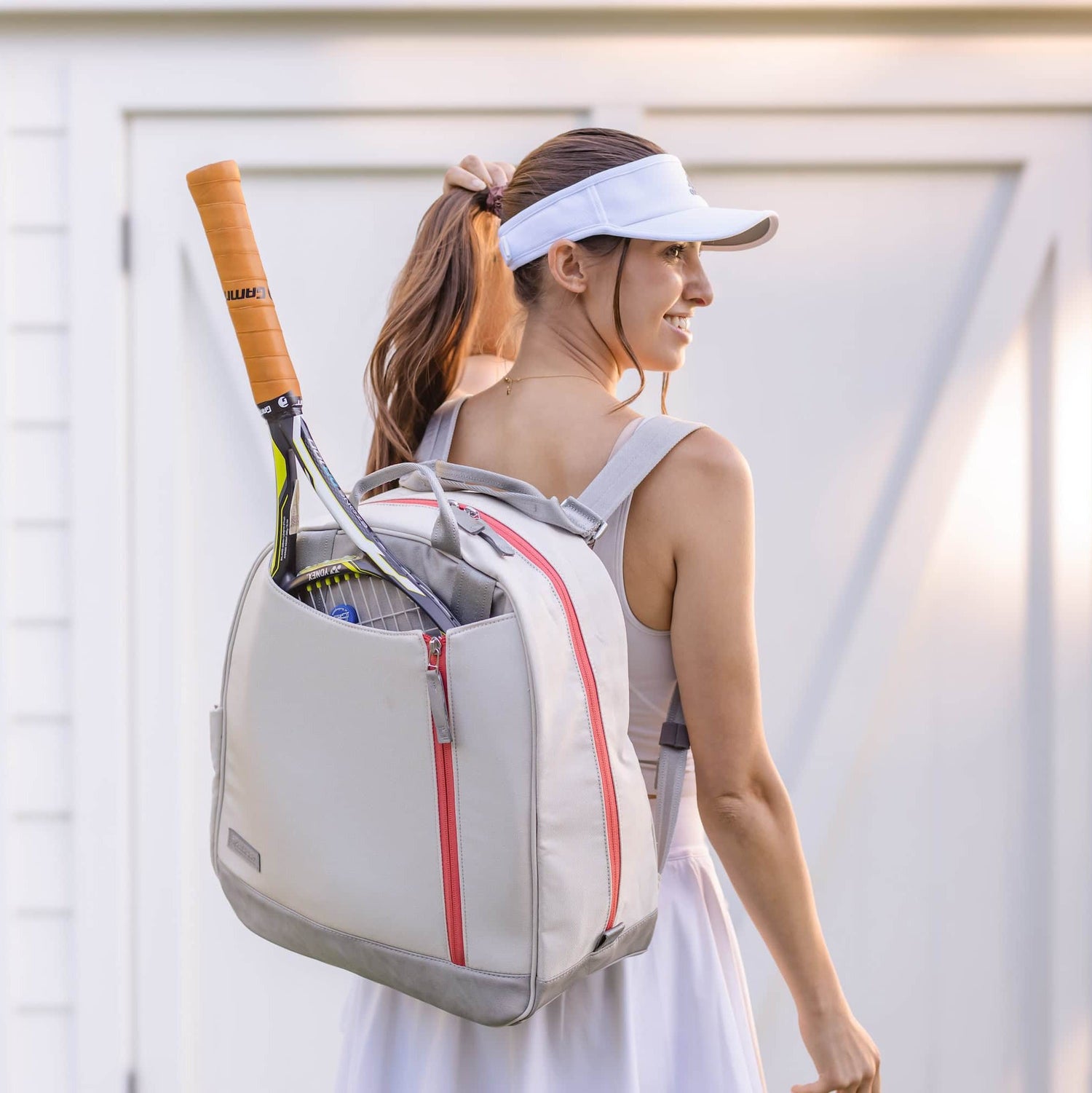 Woman with visor carries a tennis bag holding racquets, in front of a white building.