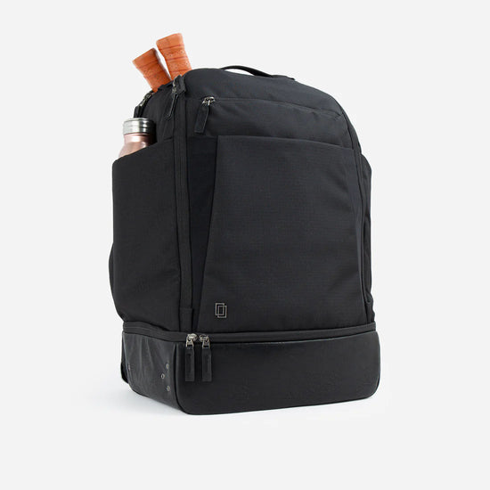 Black tennis backpack with a front pocket, water bottle, and two racquets partially visible in side pockets.