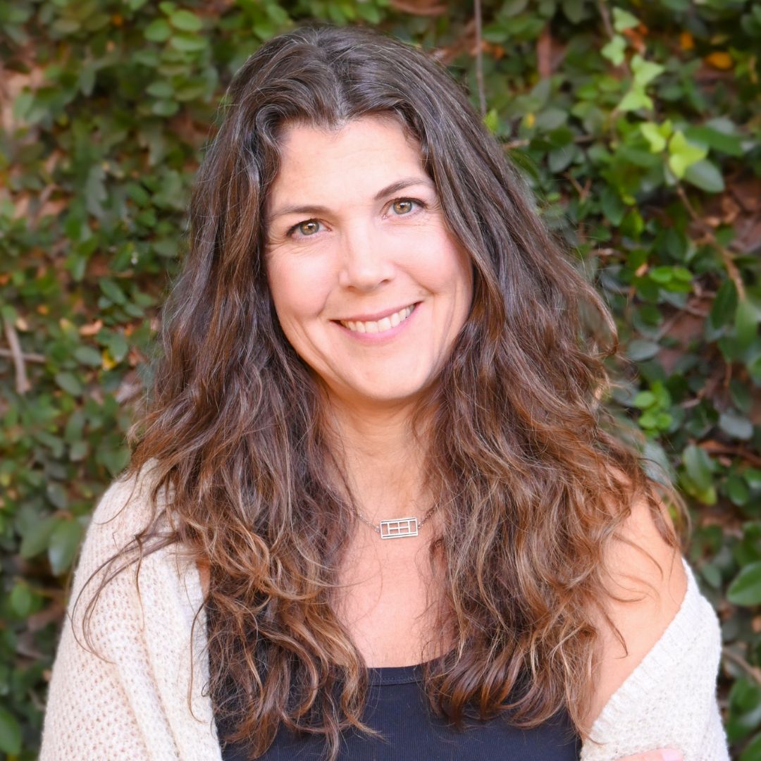 A person with long, wavy hair smiles in front of a leafy background, wearing a black top and beige shawl.