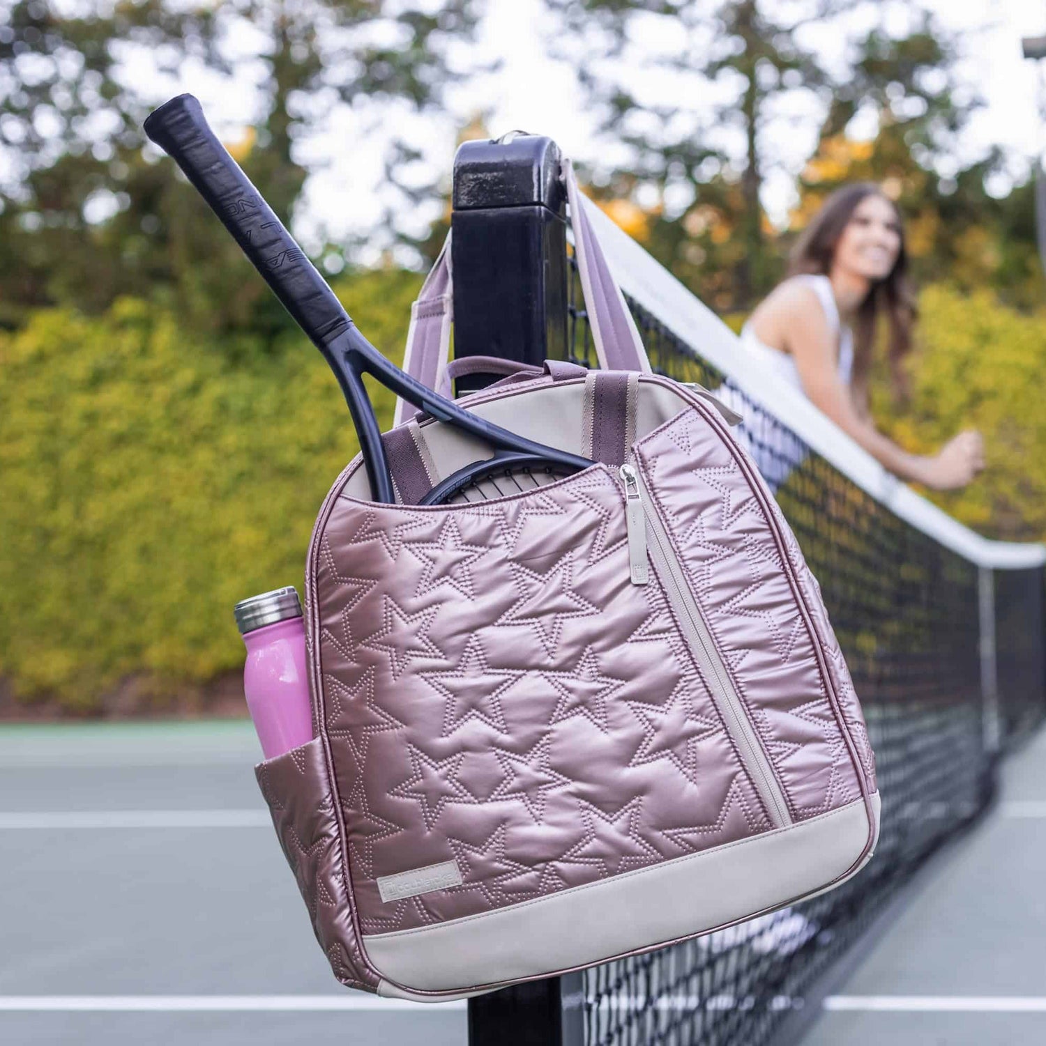Tennis bag with star pattern on net, holding a racquet and pink water bottle; a person in the background.