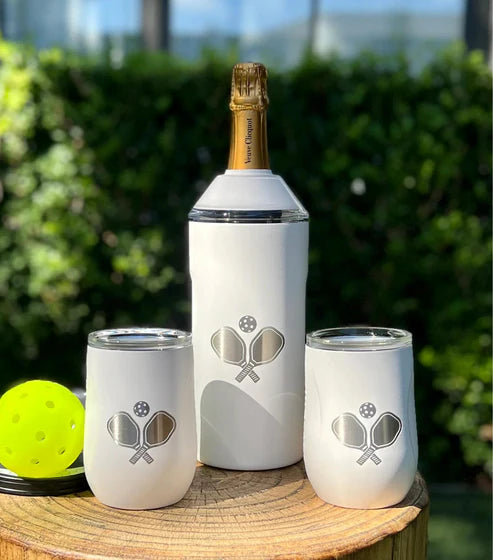 White wine bottle cooler and two tumblers with a pickleball design, next to a pickleball, outdoors.