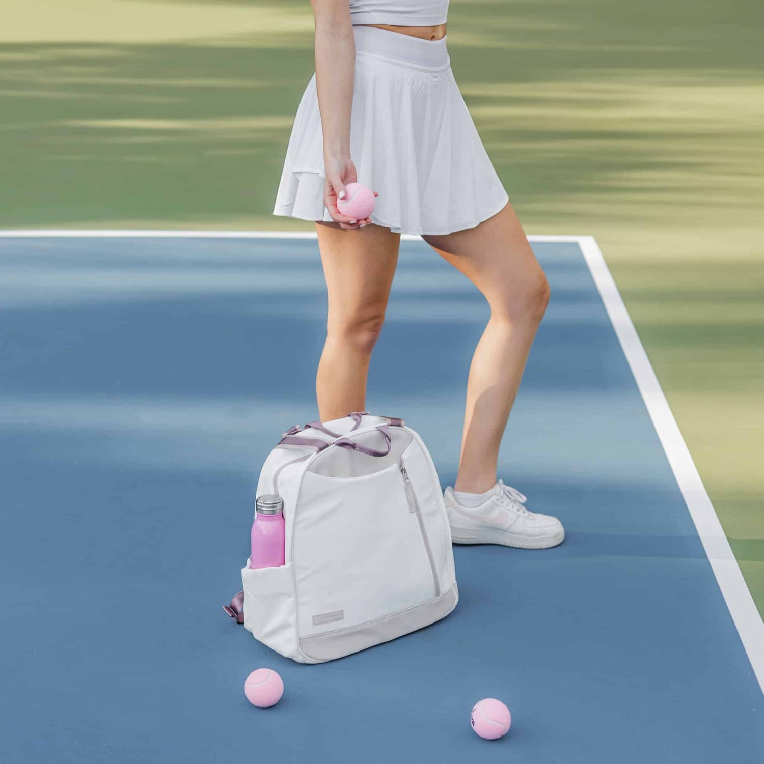 Person on a tennis court with a white tennis backpack, holding a pink ball, wearing a white skirt and sneakers.