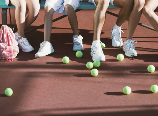 Several people sitting on a bench on a tennis court with multiple tennis balls at their feet