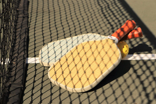 Two pickleball paddles and a pickleball in the sun in the shadow of the net