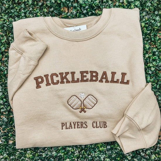 Beige sweatshirt with Pickleball Players Club and crossed paddles logo, lying on green leaves.