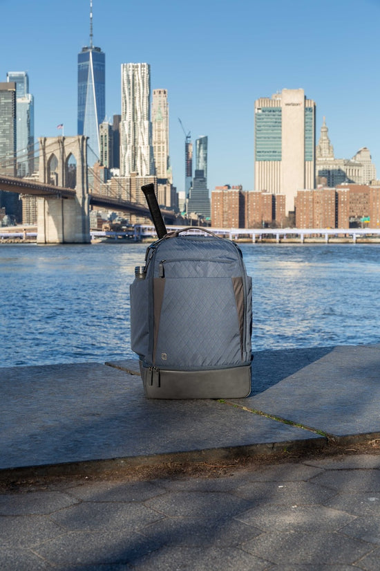 Gray tennis backpack on a concrete path with Brooklyn Bridge and city skyline in the background.