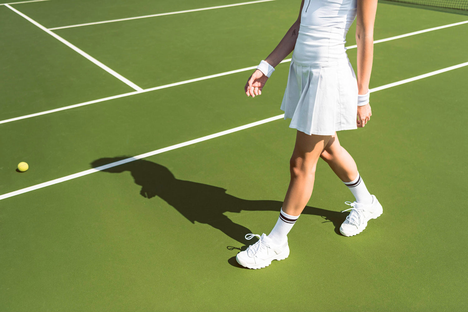 Person in a white tennis outfit walking on a green tennis court near a tennis ball.