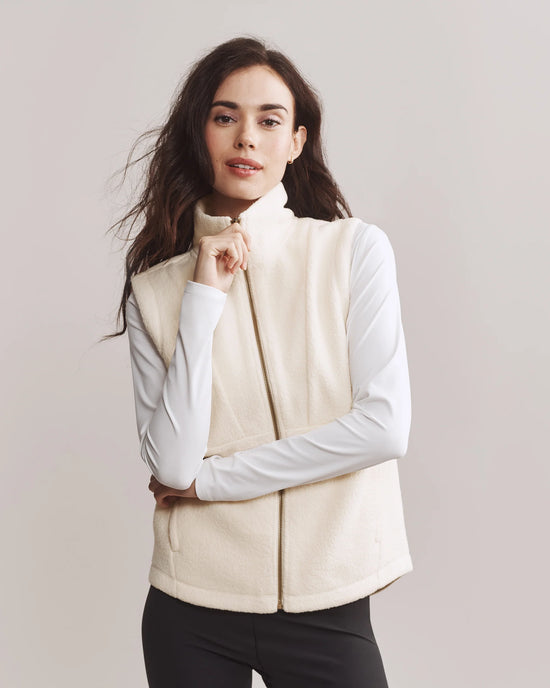 Person wearing a cream vest over a long-sleeve white shirt, standing with arms slightly crossed, against a light background.