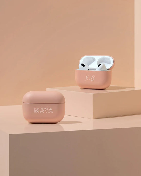 Two pink wireless earbud cases with names MAYA and K.B on a beige stepped surface. One case is open, revealing earbuds.