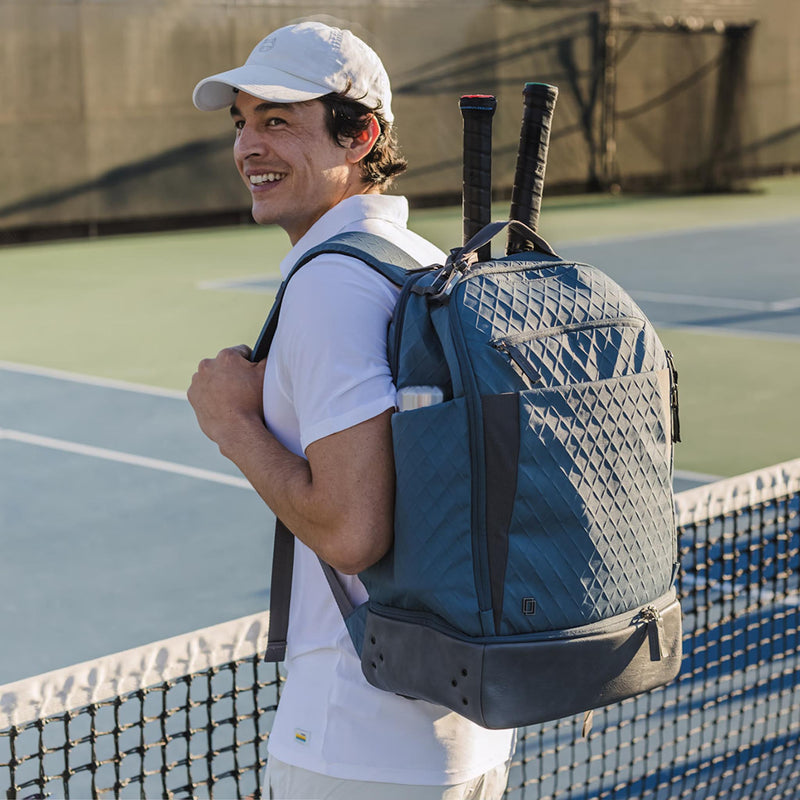 Man carrying a tennis backpack with racquets, smiling on a court during the day.