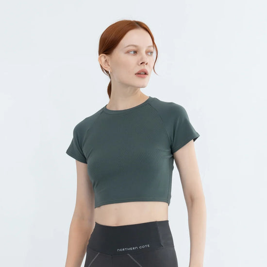 Person with red hair in a green crop top and black leggings, looking sideways against a white background.