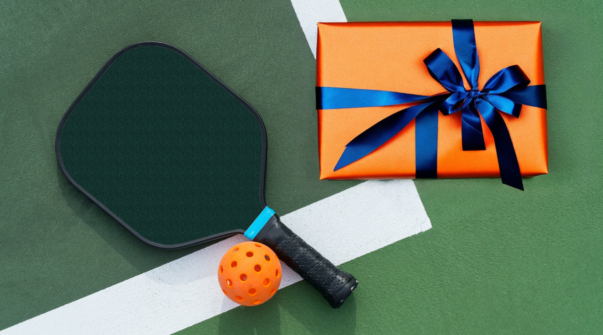 Pickleball paddle and ball next to a wrapped orange gift with a blue ribbon on a tennis court.