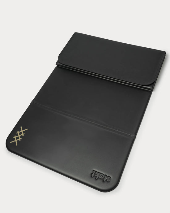A black leather laptop sleeve with a fold-over flap and a stitched logo in one corner.