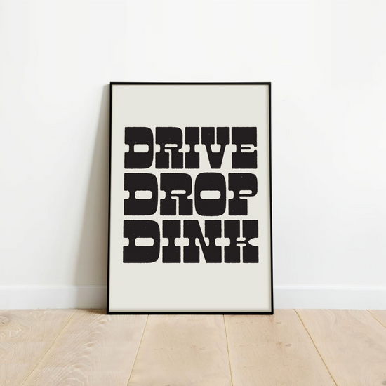 Framed poster with bold black text DRIVE DROP DINK, leaning on a white wall against a wooden floor.