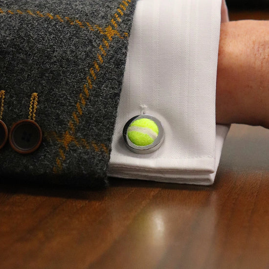Close-up of a suit sleeve with a cufflink shaped like a miniature tennis ball on a white dress shirt.
