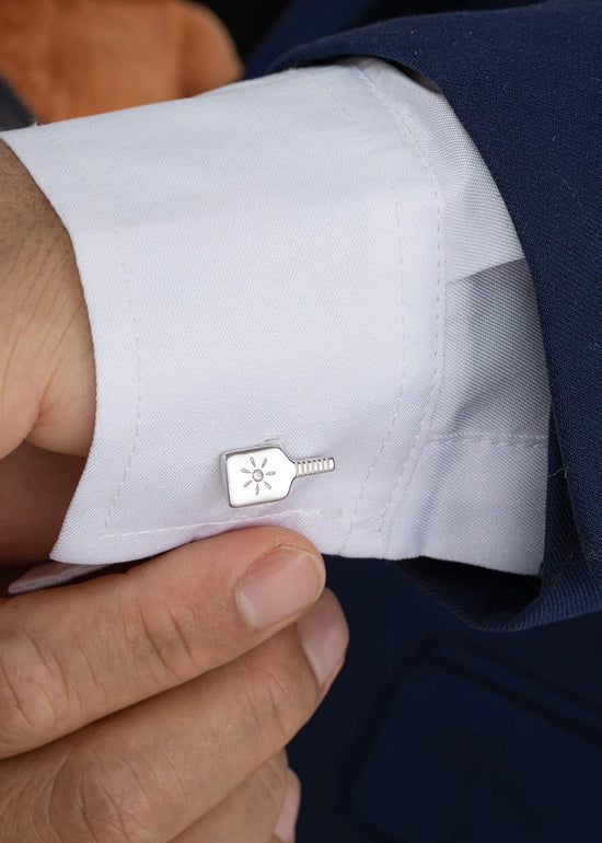 Close-up of a person adjusting a blue suit, showing a white shirt and a silver cufflink shaped like a paddle with a star.